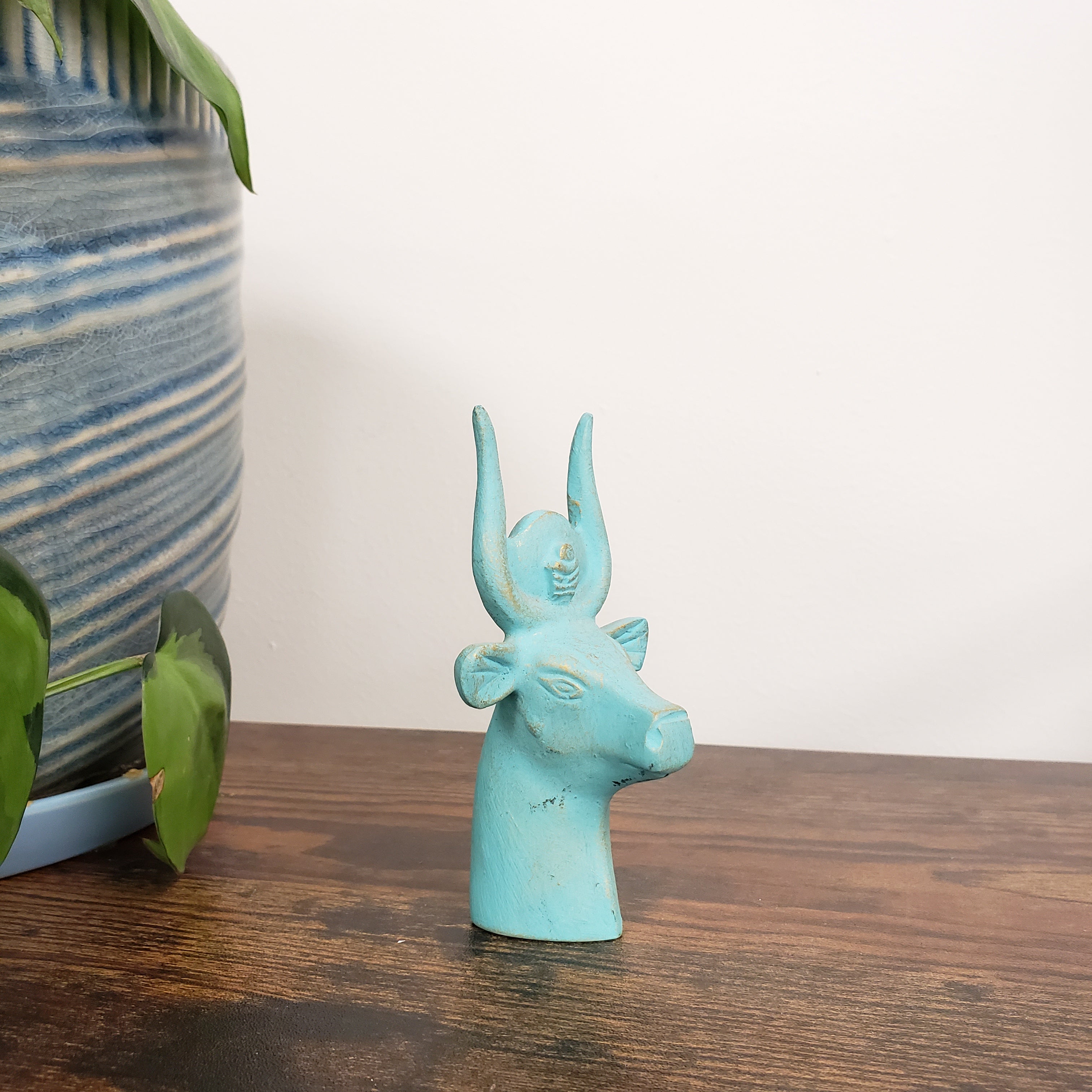 Turquoise Egyptian goddess Hathor bust statue on a wooden surface with a white wall background