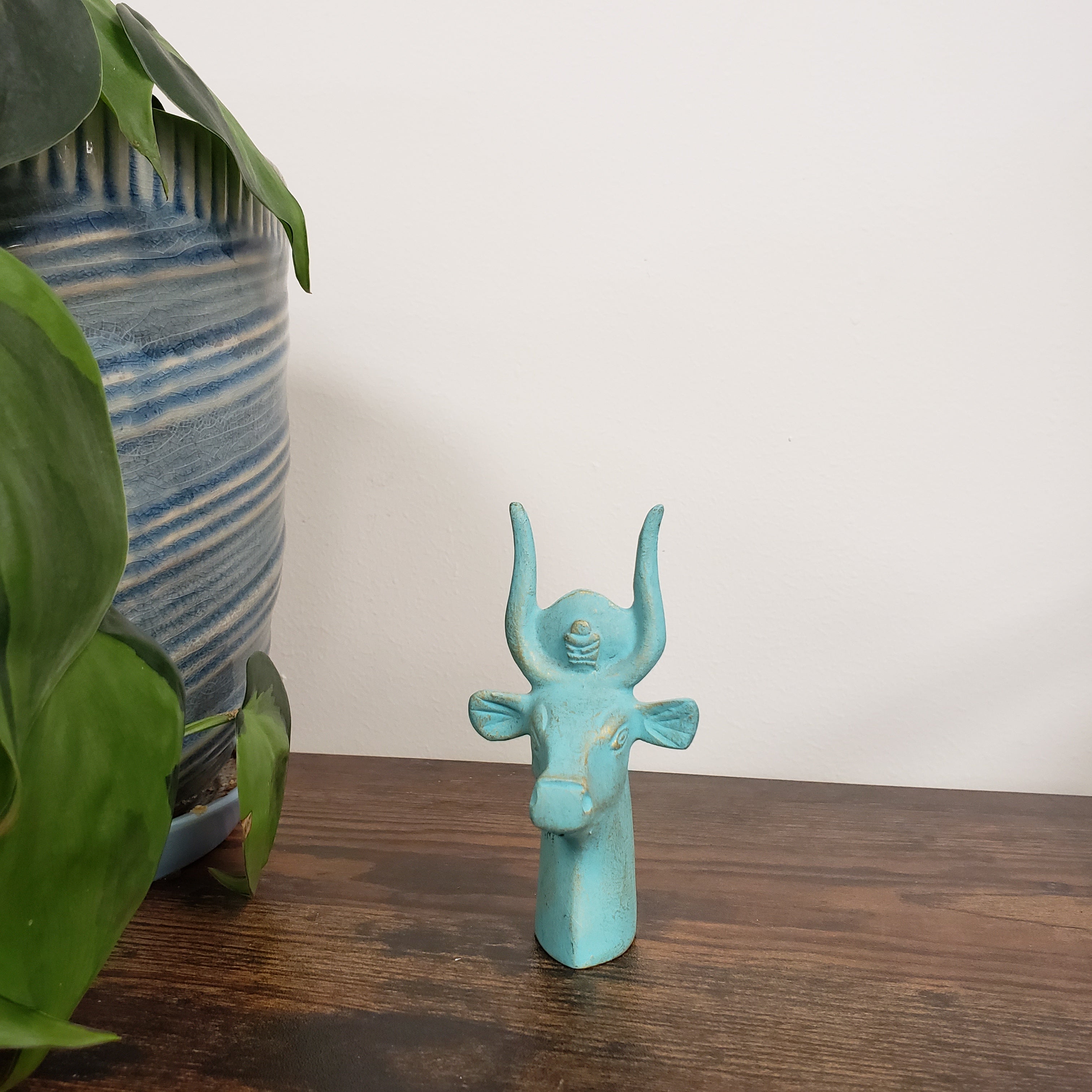 Turquoise Egyptian goddess Hathor bust statue on a wooden surface with green leaves and a striped fabric in the background
