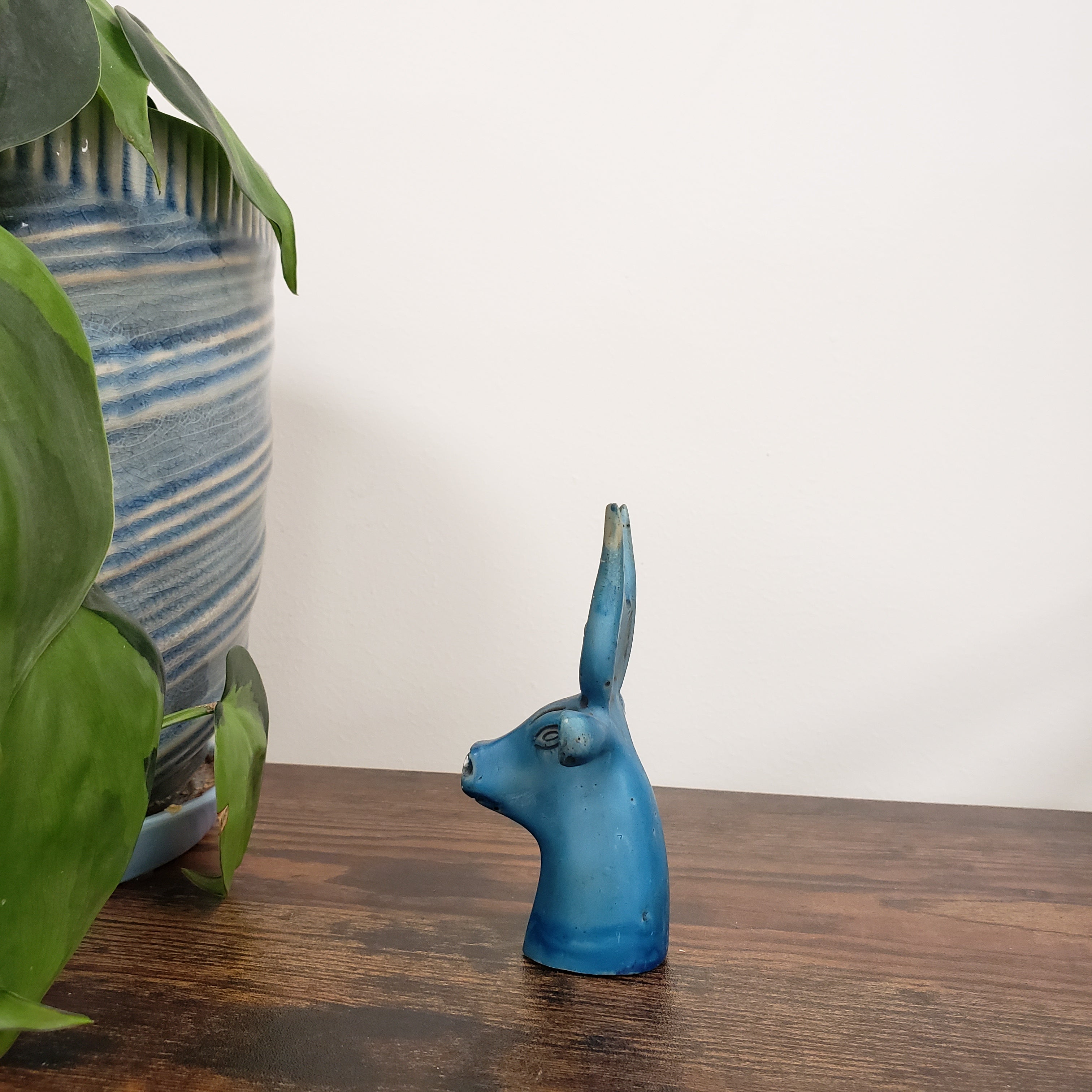 Blue Egyptian goddess Hathor bust statue on a wooden surface with plants and striped fabric in the background.
