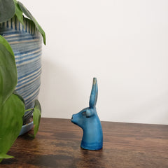 Blue Egyptian goddess Hathor bust statue on a wooden surface with plants and striped fabric in the background.