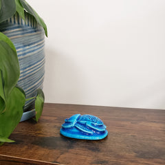 Blue ceramic scarab beetle on a wooden surface with green leaves in the foreground