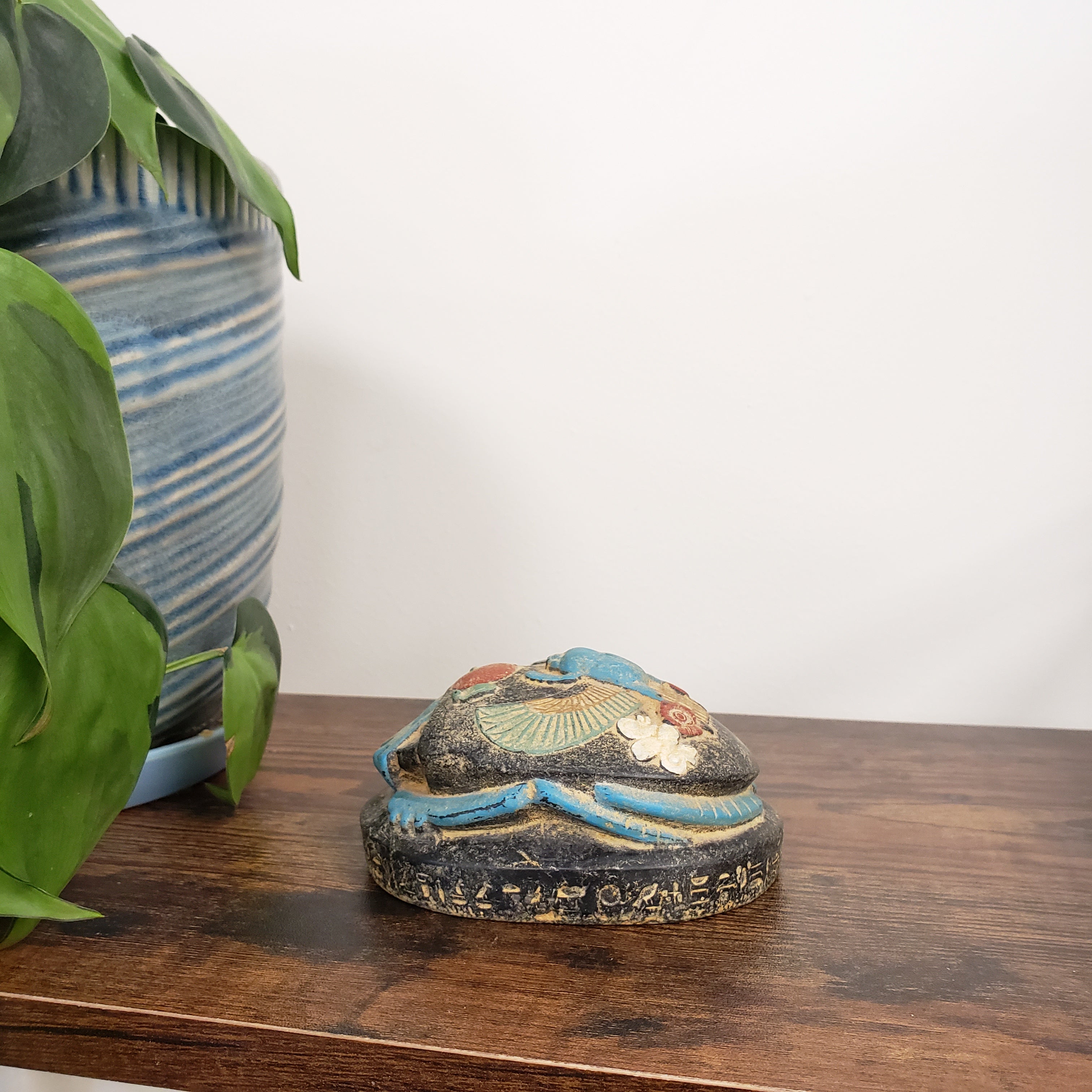 Decorative Egyptian scarab statue on a wooden panel with plants and striped fabric in the background