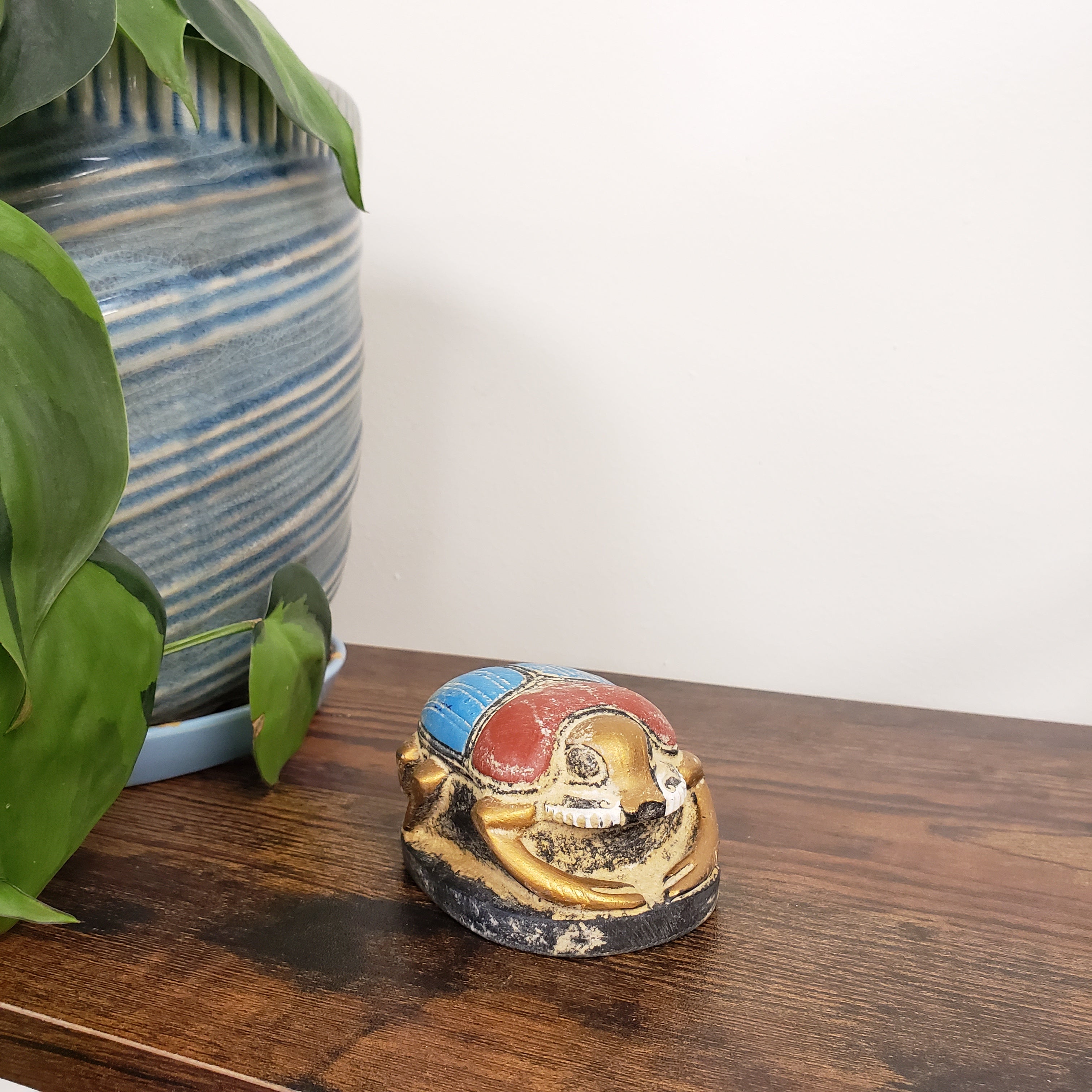 Decorative Egyptian scarab statue on a wooden surface with plants and a striped pillow in the background