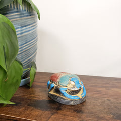 Decorative Egyptian scarab statue with blue and gold accents on a wooden surface with green leaves in the foreground.