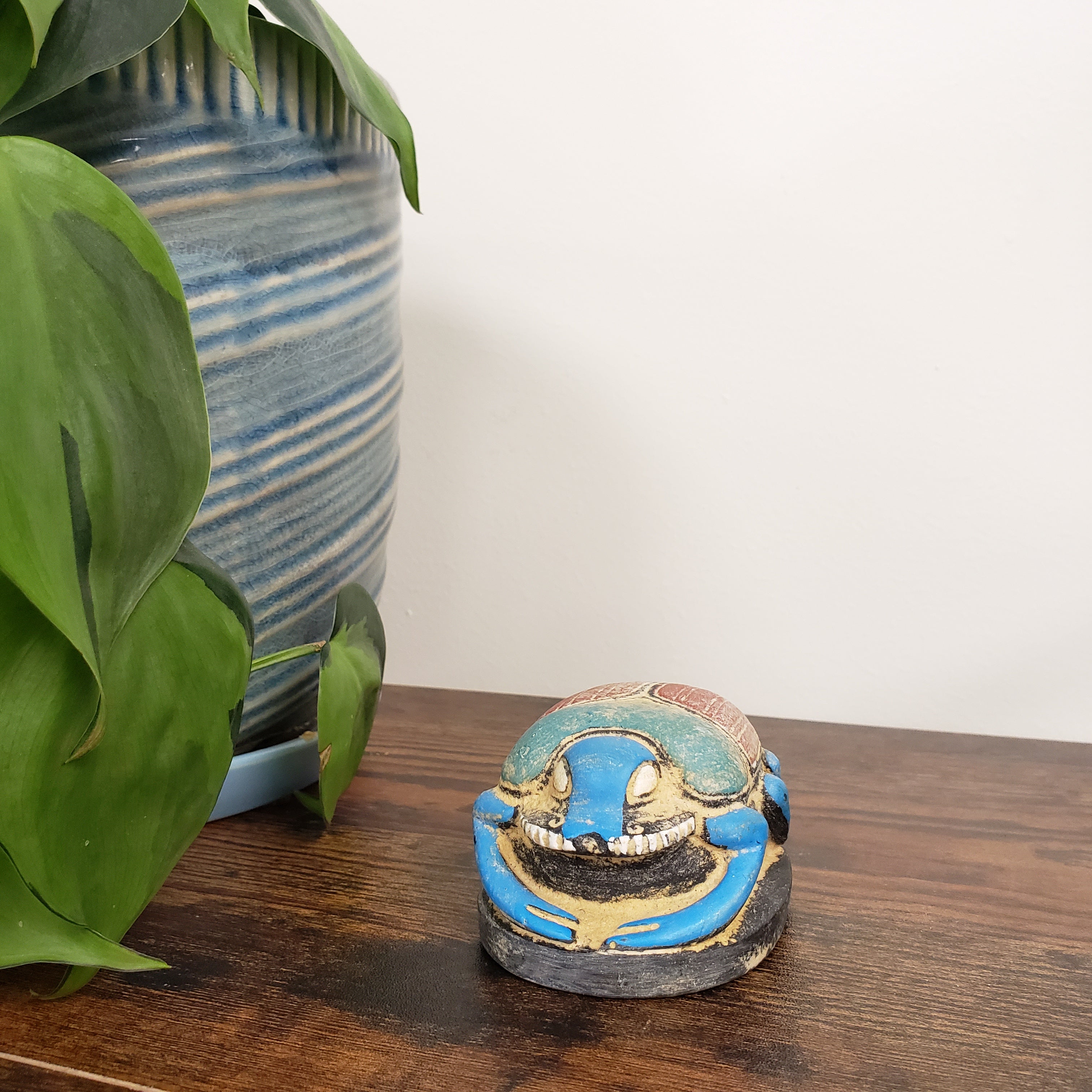 Decorative blue and gold Egyptian scarab statue on a wooden surface with a plant and ceramic pot in the background.