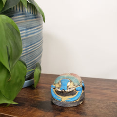 Decorative blue and gold Egyptian scarab statue on a wooden surface with a plant and ceramic pot in the background.