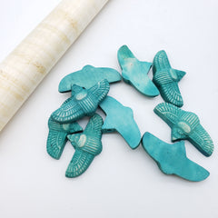 Six turquoise-colored wooden beads shaped like stylized birds, arranged on a white surface next to a rolled piece of natural beige shell or paper.