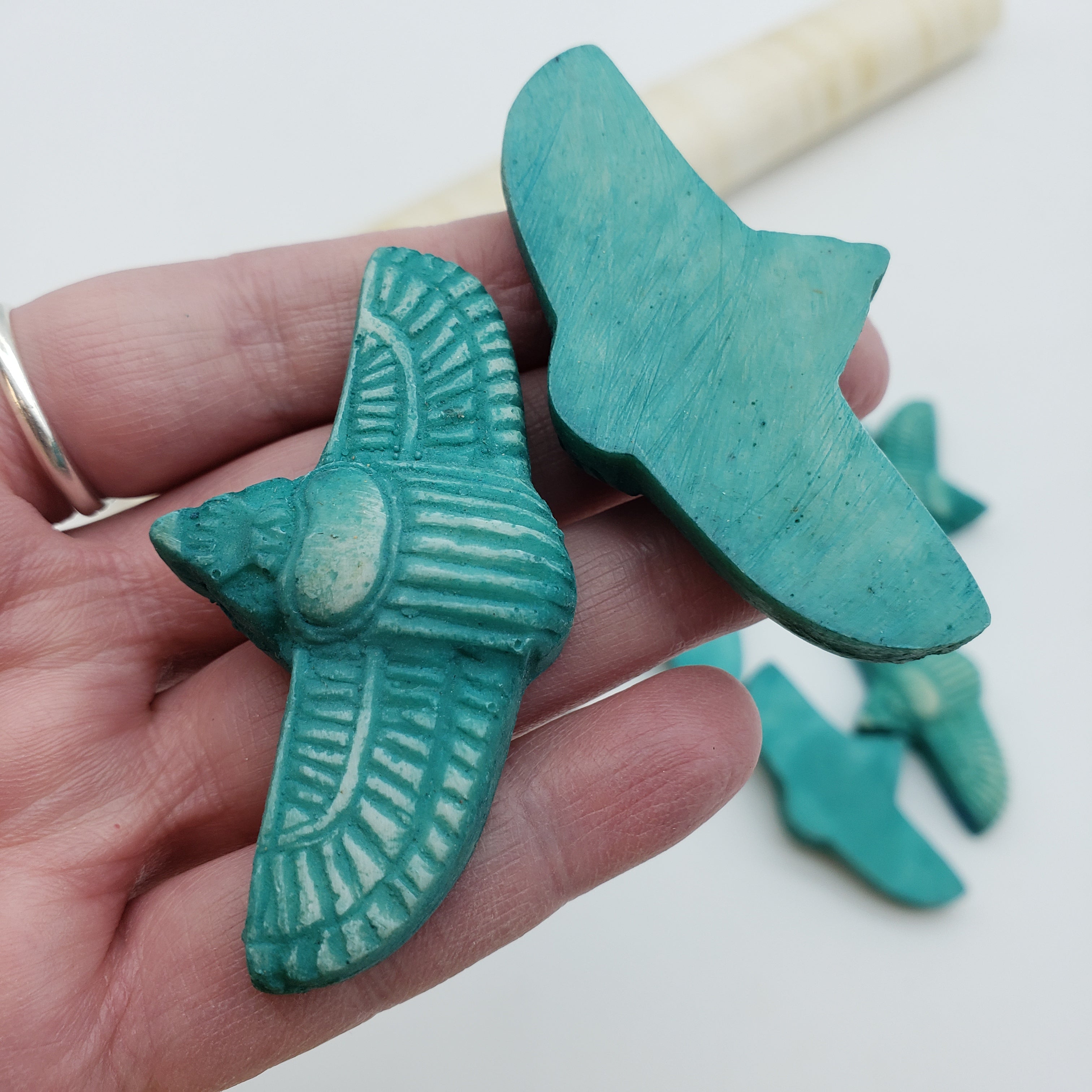 A hand holds two turquoise-colored carved Egyptian scarab amulets with a few more pieces blurred in the background on a white surface.