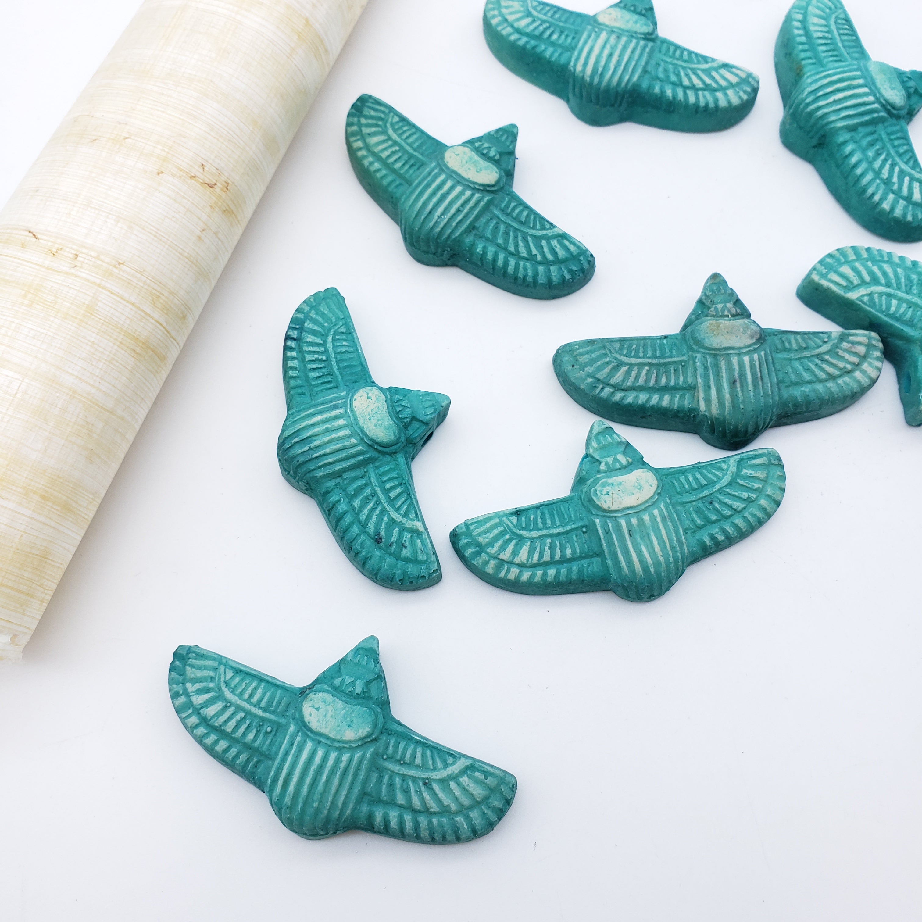 Turquoise scarab-shaped beads with a carved textured pattern, arranged on a white surface next to a rolled seashell.