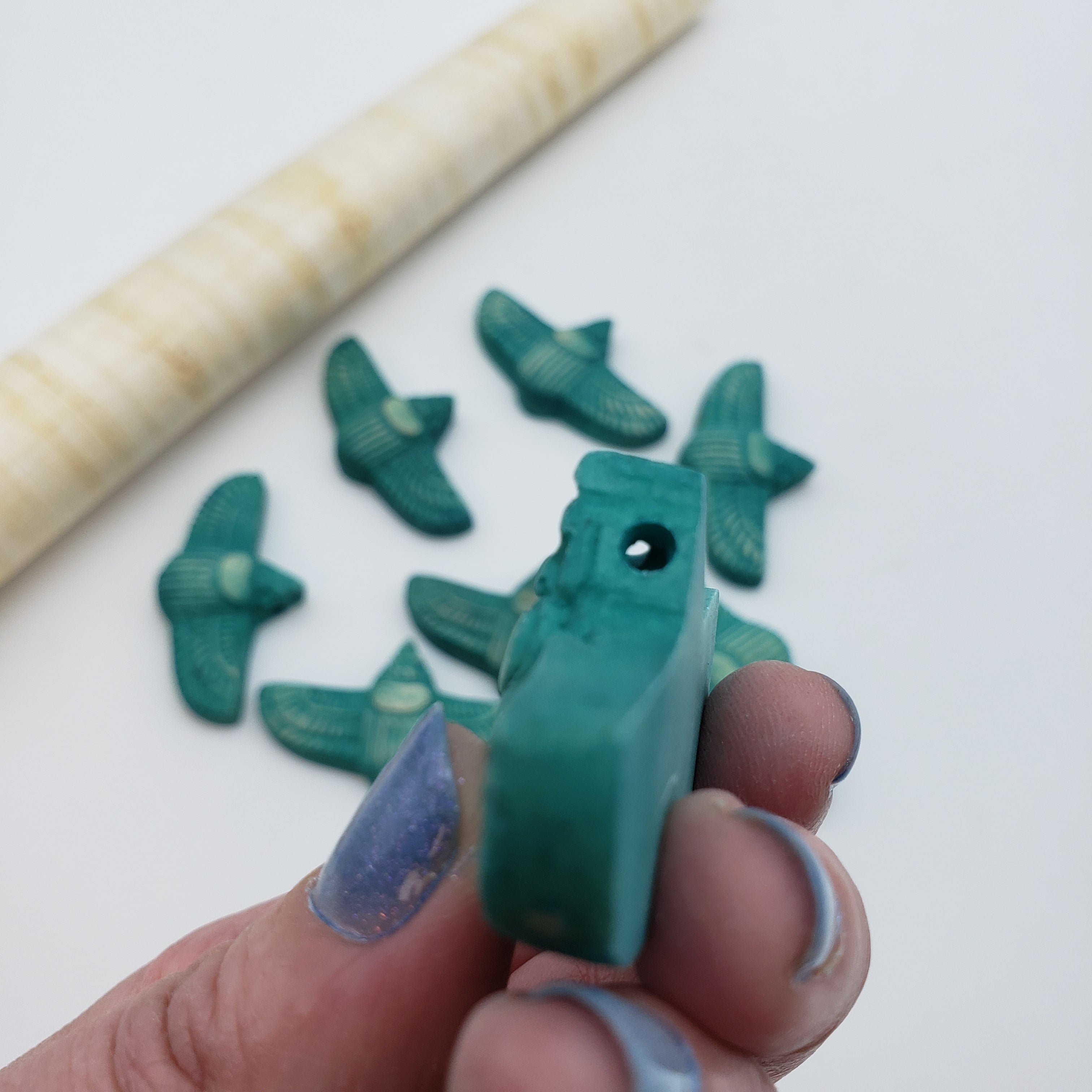 A hand holds a small teal-green carved scarab-shaped bead or pendant, with several matching beads scattered in the background and a rolled papyrus prop on a white surface.