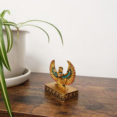 Decorative statue with Egyptian-style design with a gold and blue design on a wooden surface with a plant in the foreground.