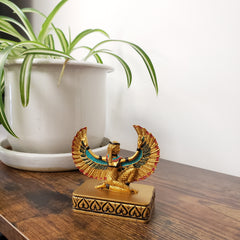Decorative statue with Egyptian-style design on a wooden surface next to a plant.