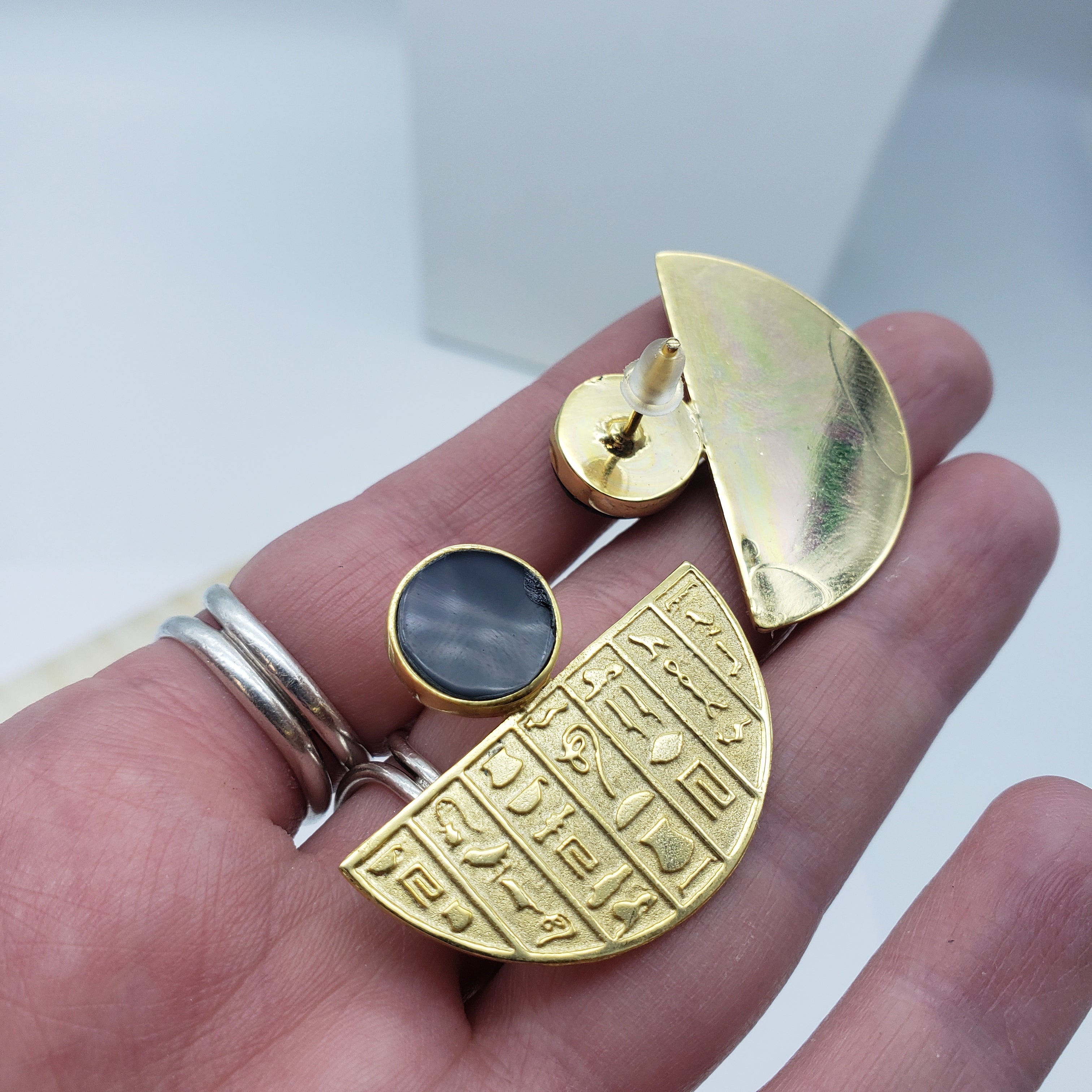 Gold earring with hieroglyphic design held in a hand against a neutral background