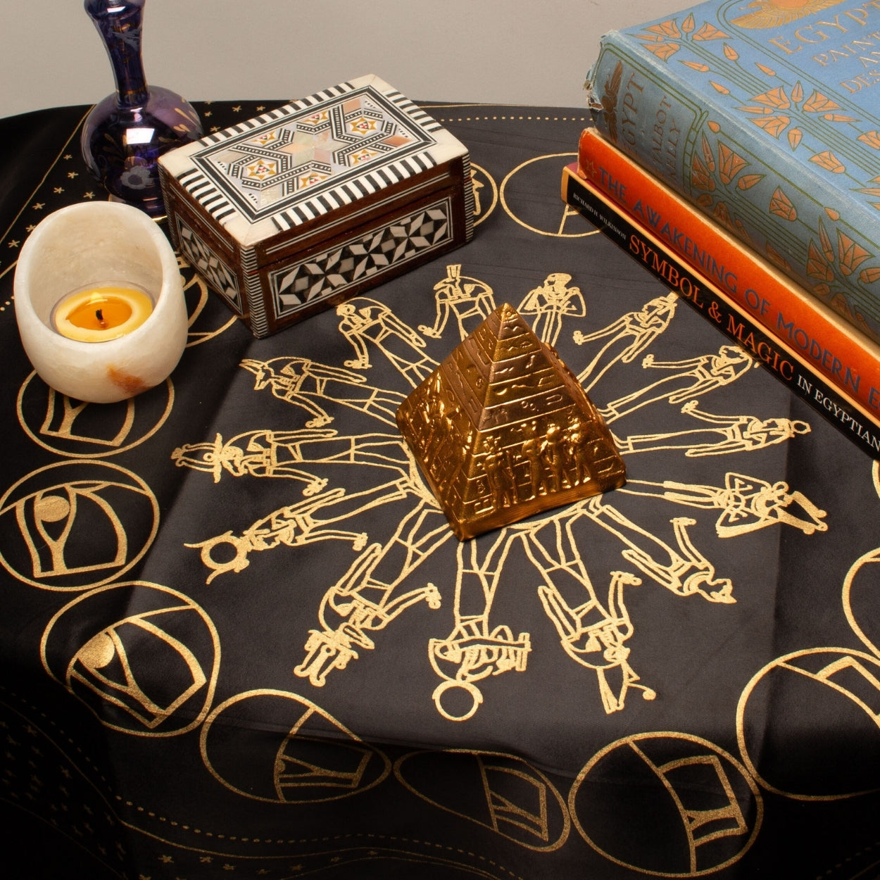 Decorative table setting with Egyptian-themed items including a candle, books, and a small pyramid model on a black cloth with gold symbols.