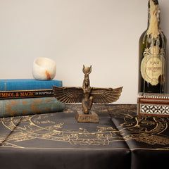 Bronze statue of a winged Isis figure on a table with books and a bottle.