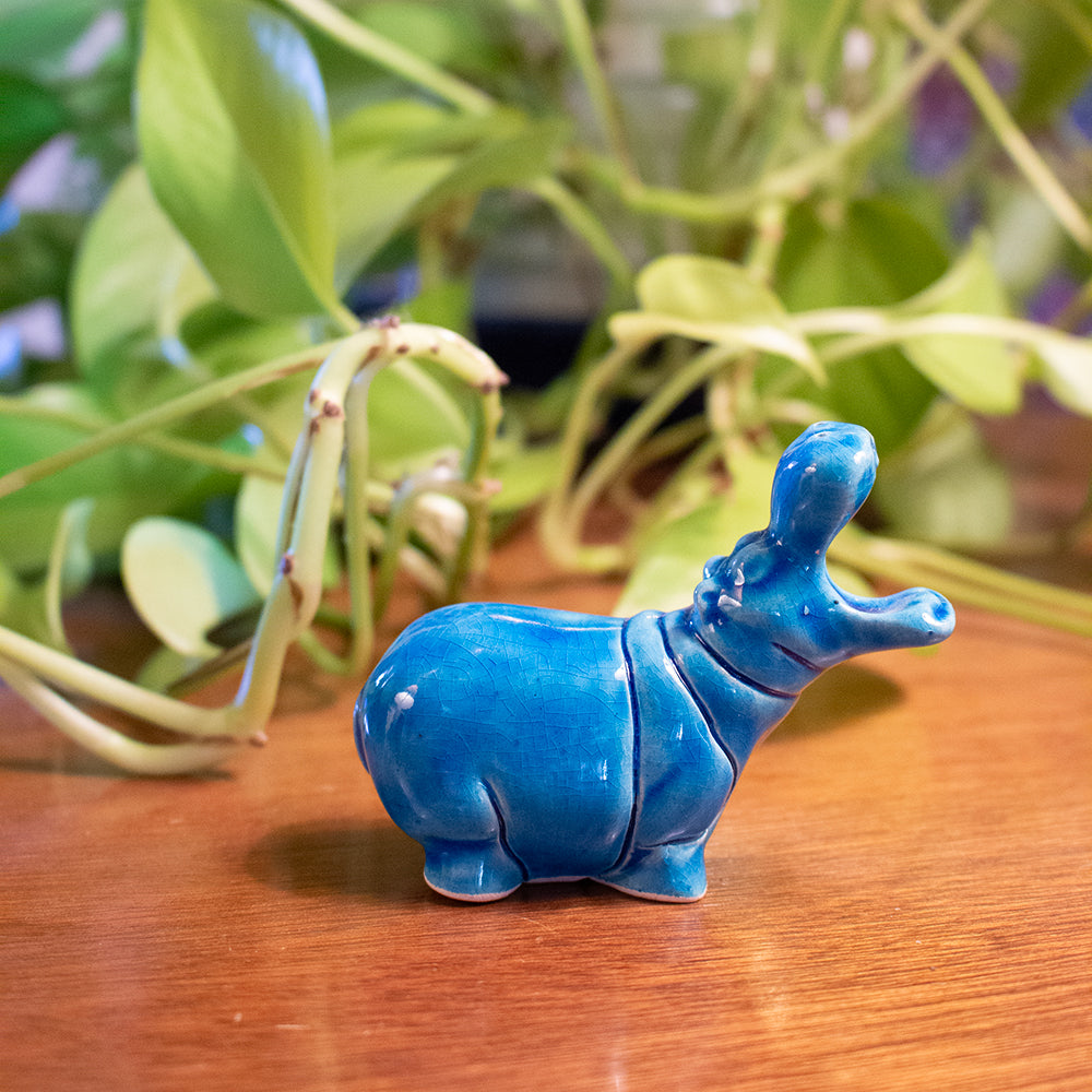 Blue ceramic hippo figurine on a wooden surface with green plants in the background