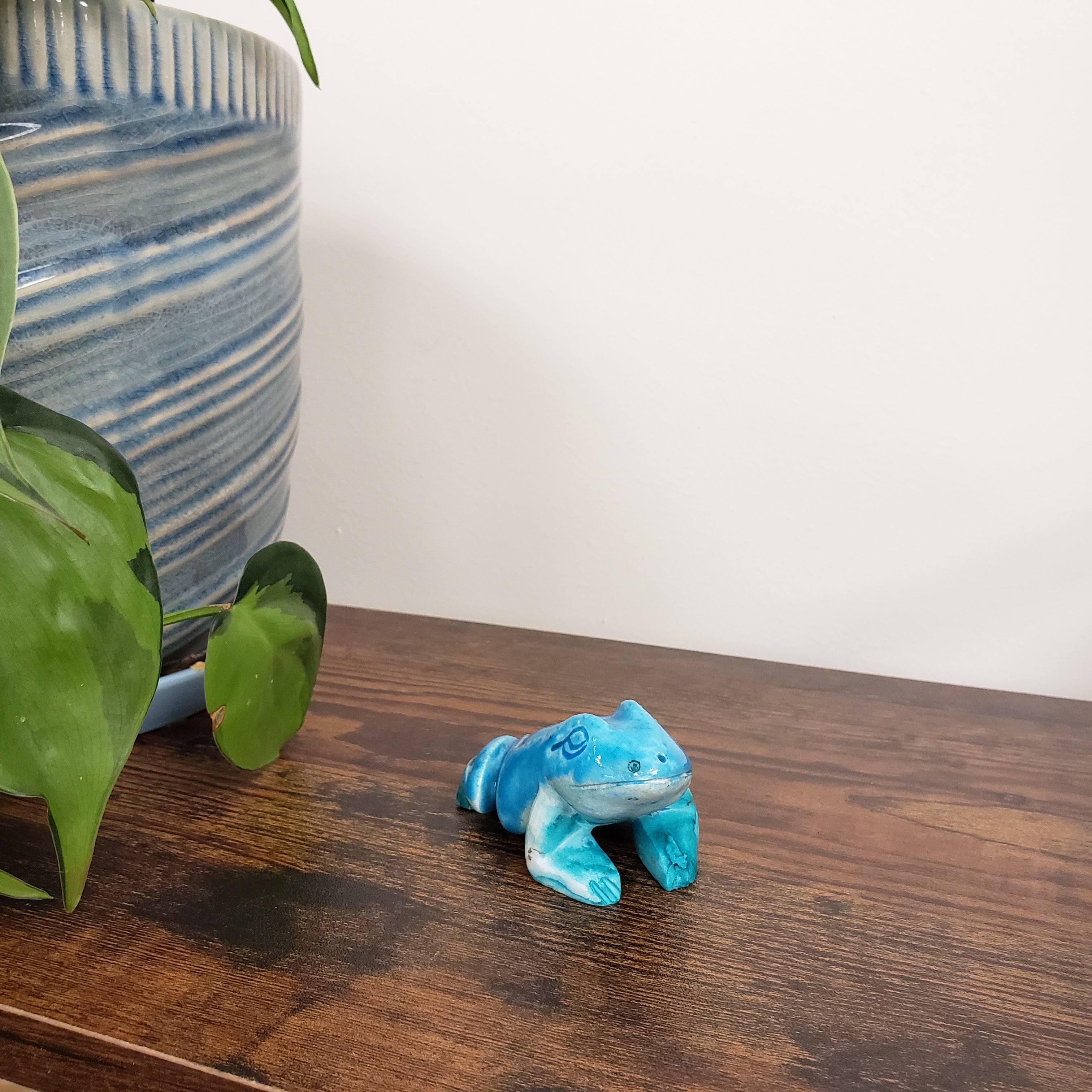 Blue ceramic frog figurine on a wooden surface next to a plant and ceramic pot.