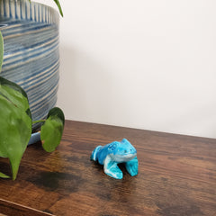 Blue ceramic frog figurine on a wooden surface next to a plant and ceramic pot.