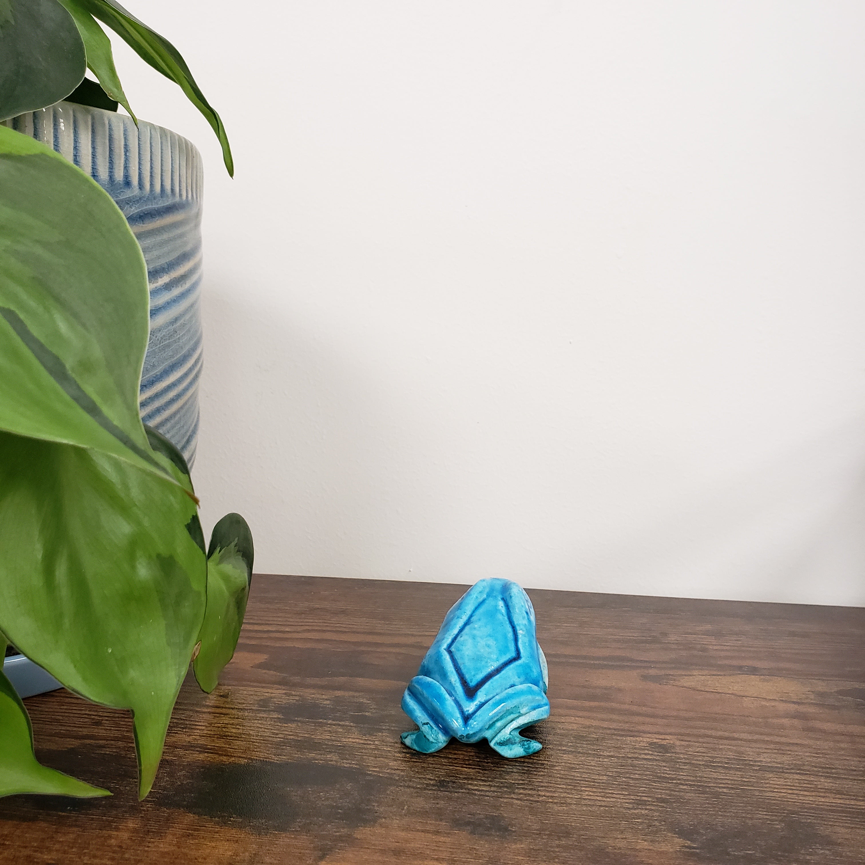 Blue decorative ceramic frog figurine item on a wooden surface with green leaves and a striped cushion in the background