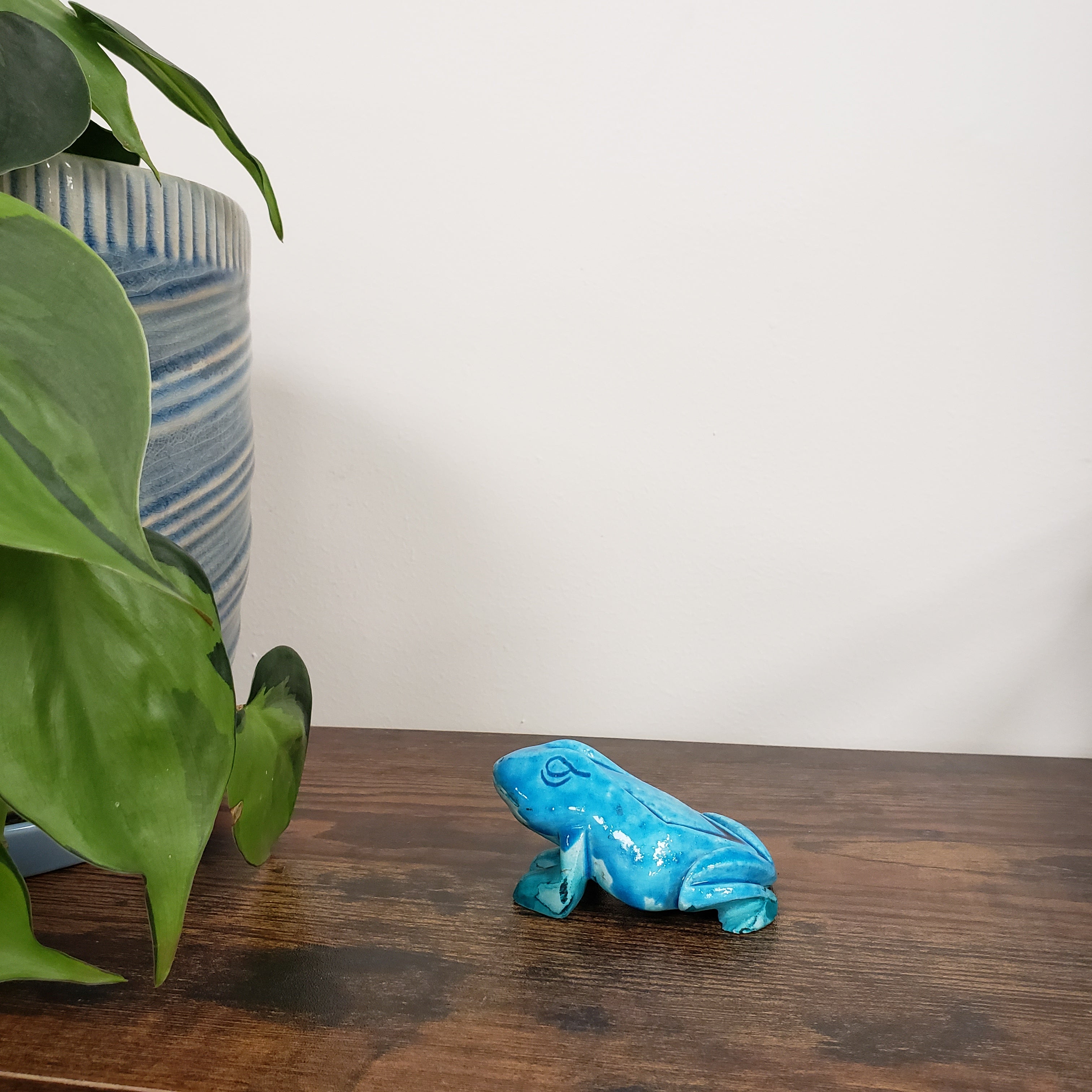Blue ceramic frog figurine on a wooden surface with green leaves and striped fabric in the background.