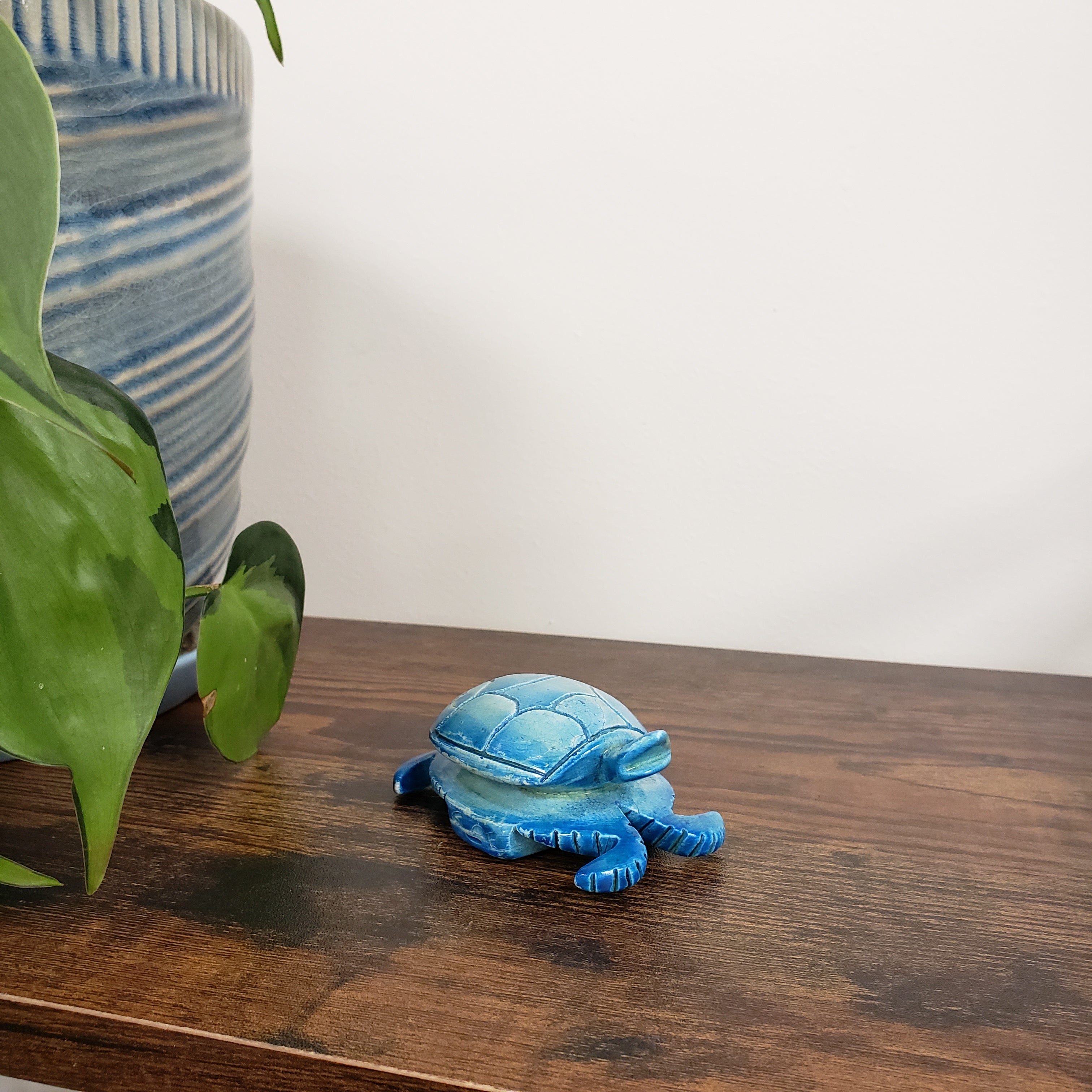 Blue ceramic turtle figurine on a wooden surface with a striped cushion and plant in the background.