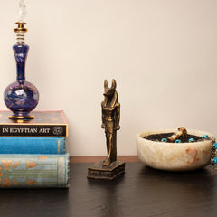 A bronze Anubis statue stands on a dark wooden table between a stack of vintage books and a white marble bowl with turquoise beads, against a plain beige wall.