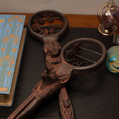 An ornate dark bronze-finished sistrum object with Egyptian-style relief carvings, displayed on a dark tabletop next to a blue patterned book and other decorative objects.