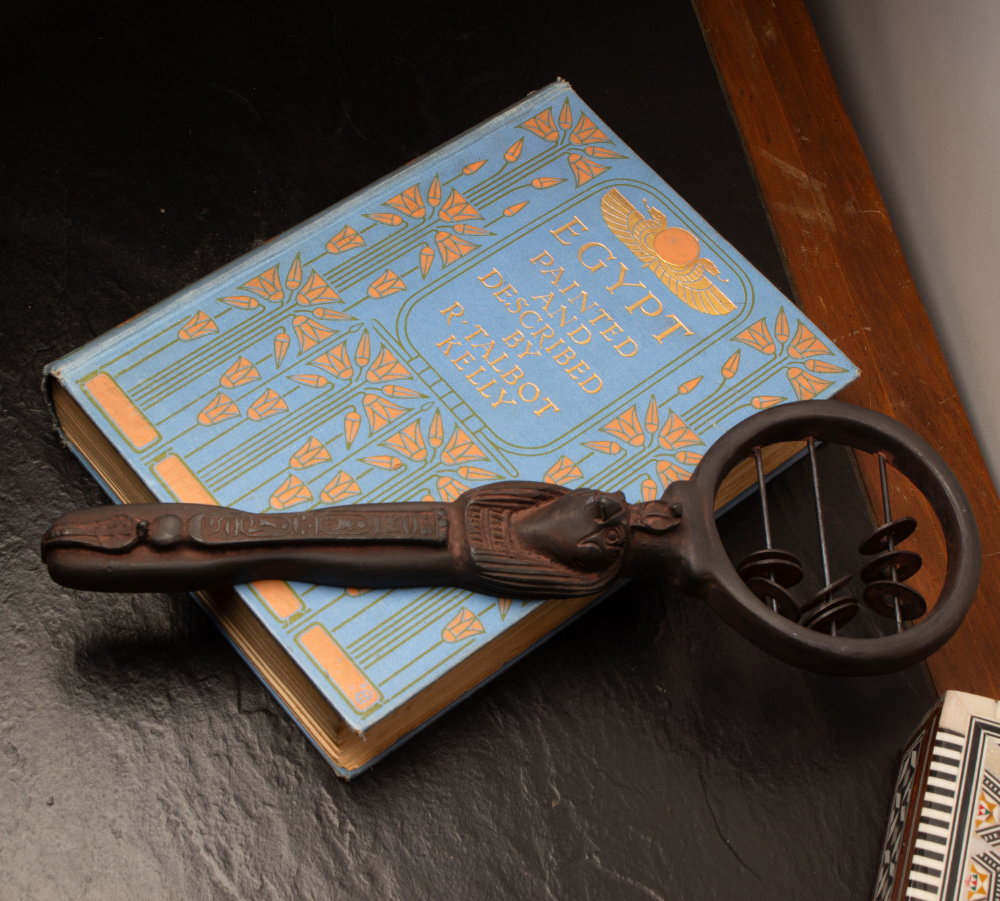 An ancient Egyptian sistrum rattle, resting diagonally on top of a light blue book titled 'The Egypt Unearthed' on a dark slate surface.