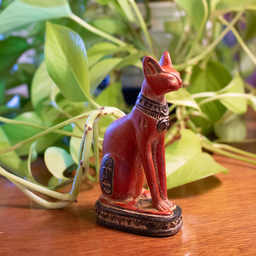 Red cat figurine with hieroglyphics on a wooden surface with green plants in the background