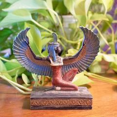 Statue of a winged figure on a wooden base with green plants in the background