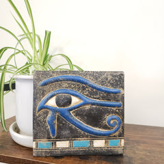 Decorative wall art with blue eye design on a wooden surface with a plant in the background