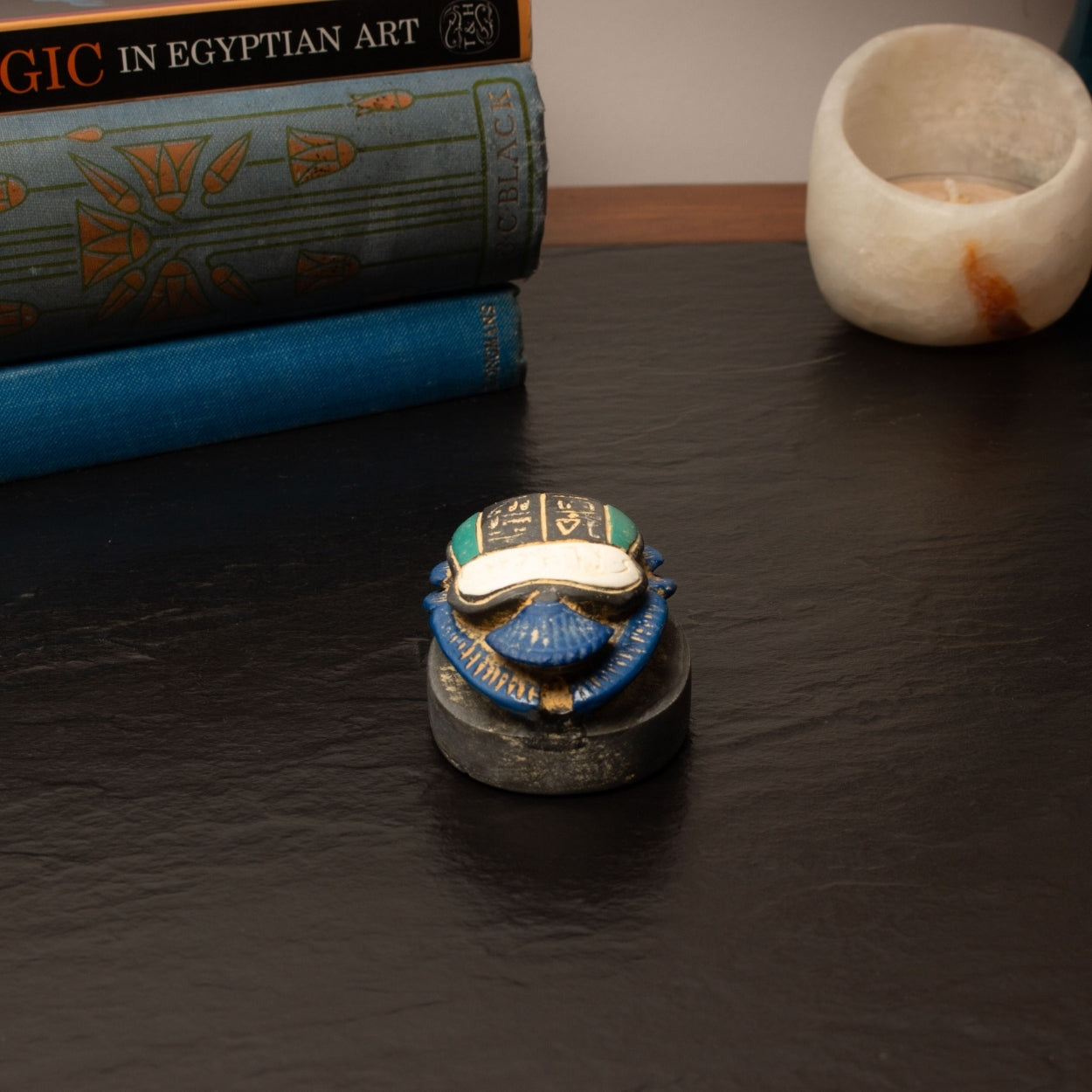 Decorative scarab with blue and gold details on a dark surface, with books and a small bowl in the background.
