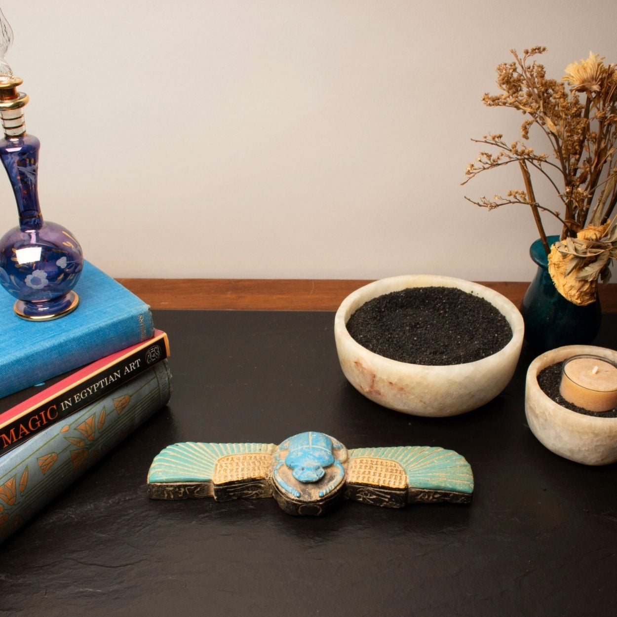 Decorative items including a blue lamp, books, a bowl with soil, a scarab, and a candle on a dark surface.