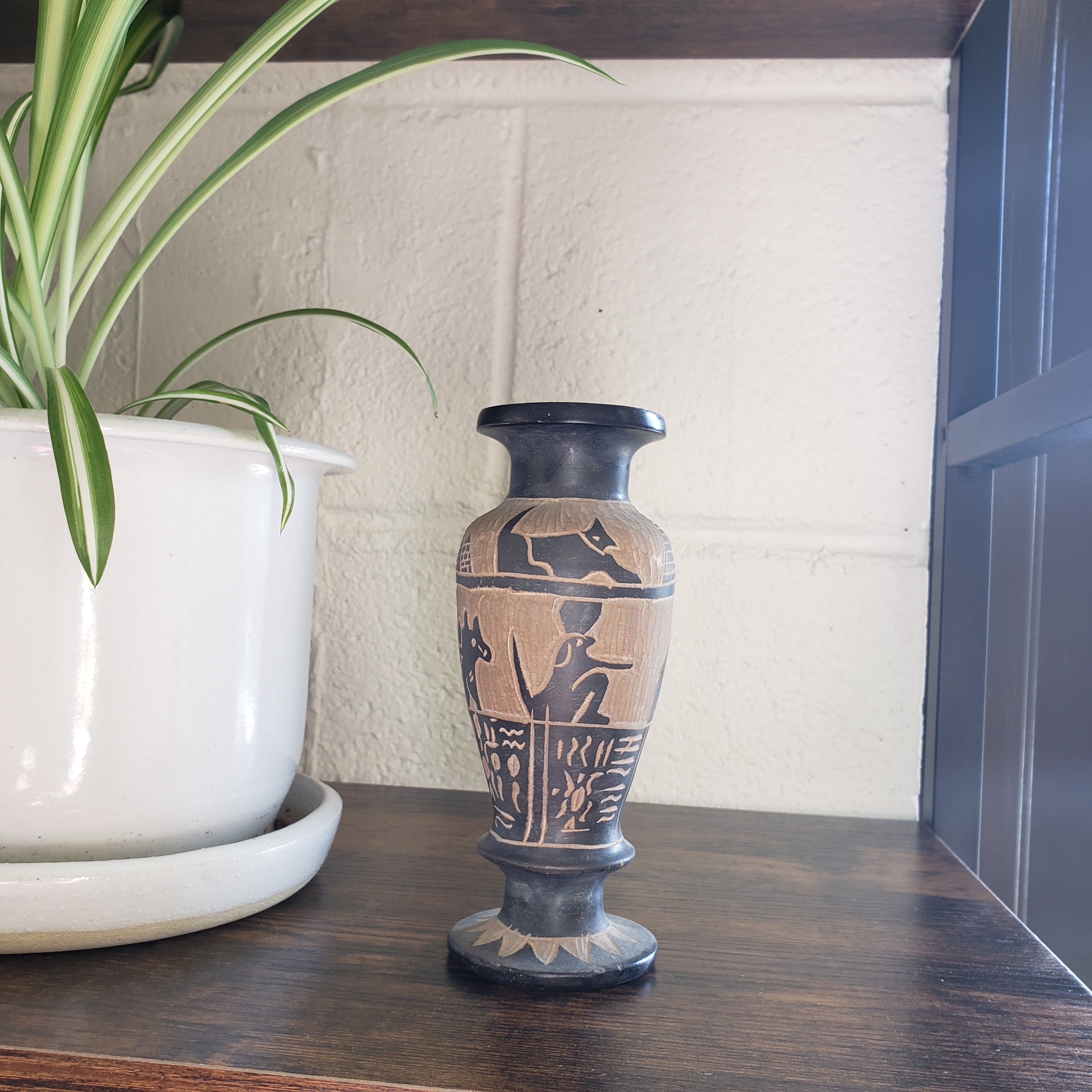 Decorative vase with hieroglyphics on a wooden shelf with a plant in the foreground.