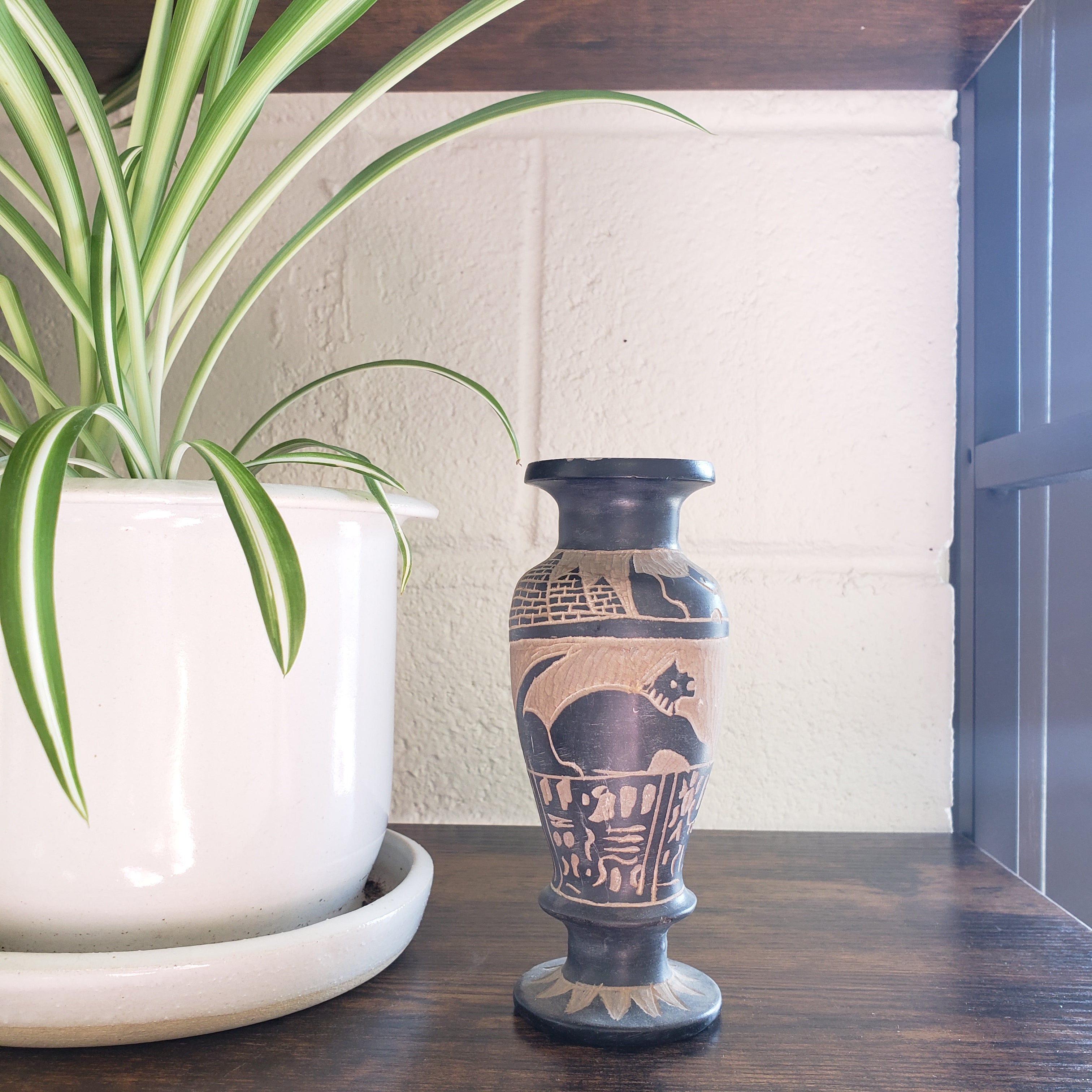 Decorative vase with intricate patterns on a wooden shelf with a plant in the foreground.