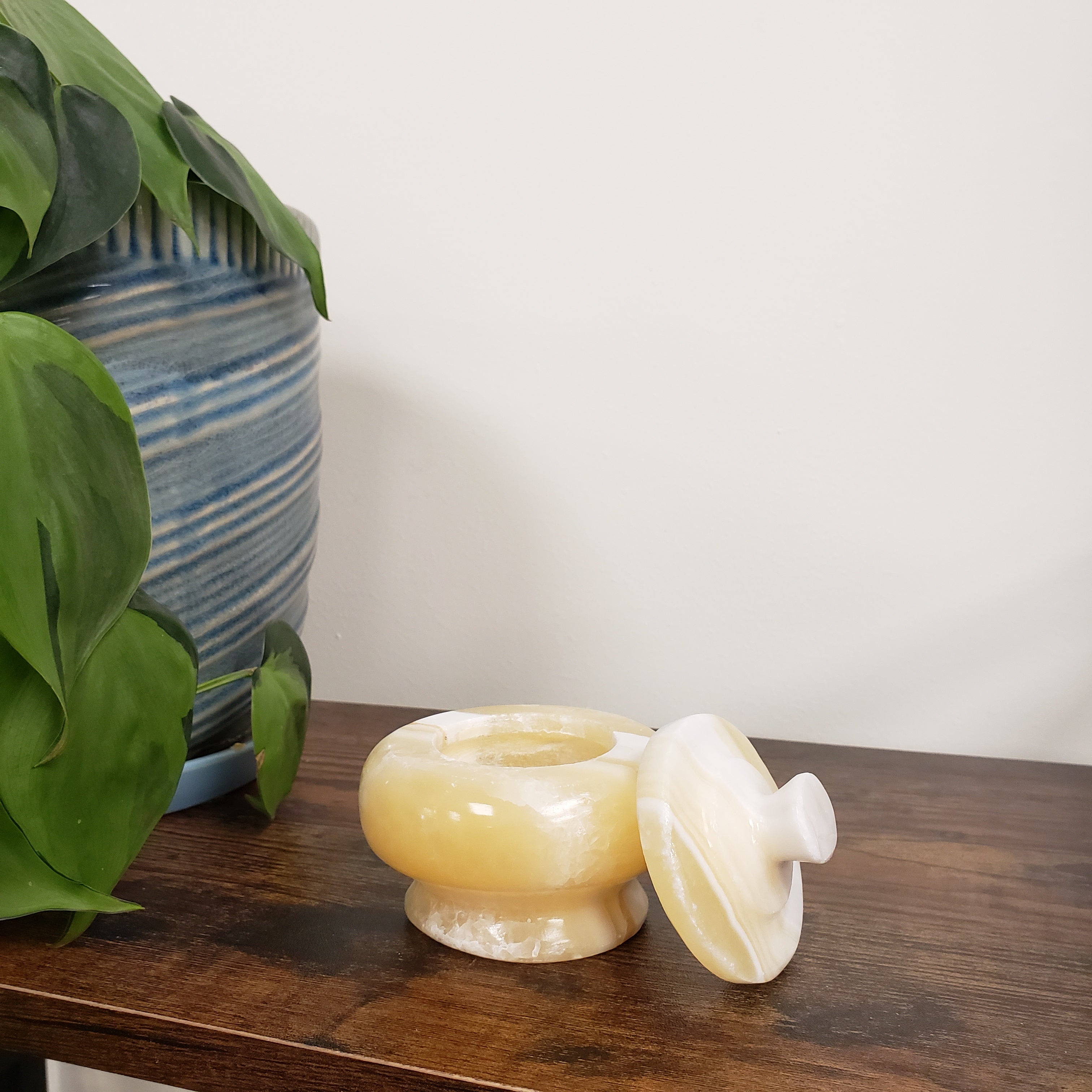 Decorative alabaster jar on a wooden surface with a plant and striped fabric in the background.