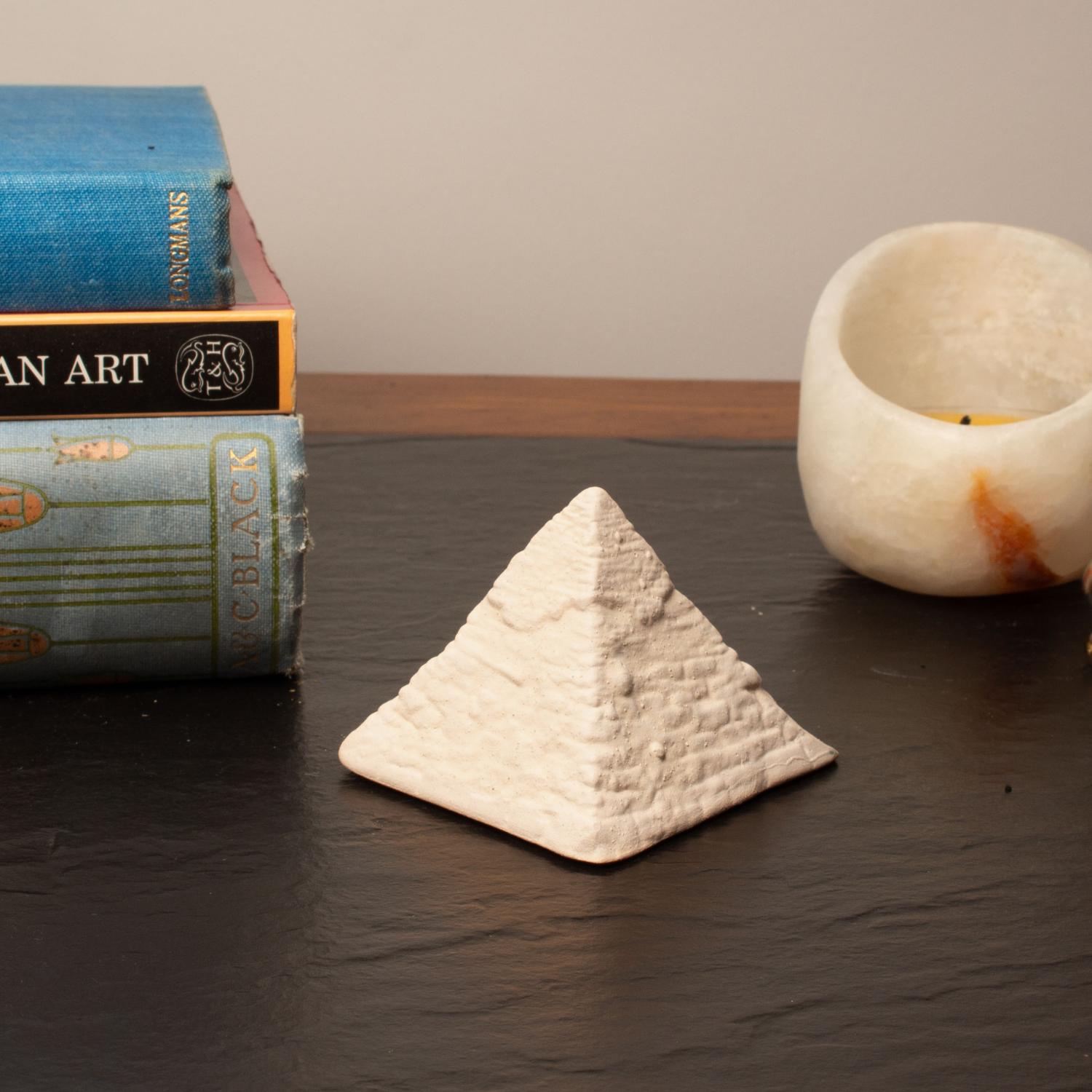 A small textured white stone pyramid figurine sits on a dark tabletop, styled with vintage books on the left and a white marble candle holder on the right.