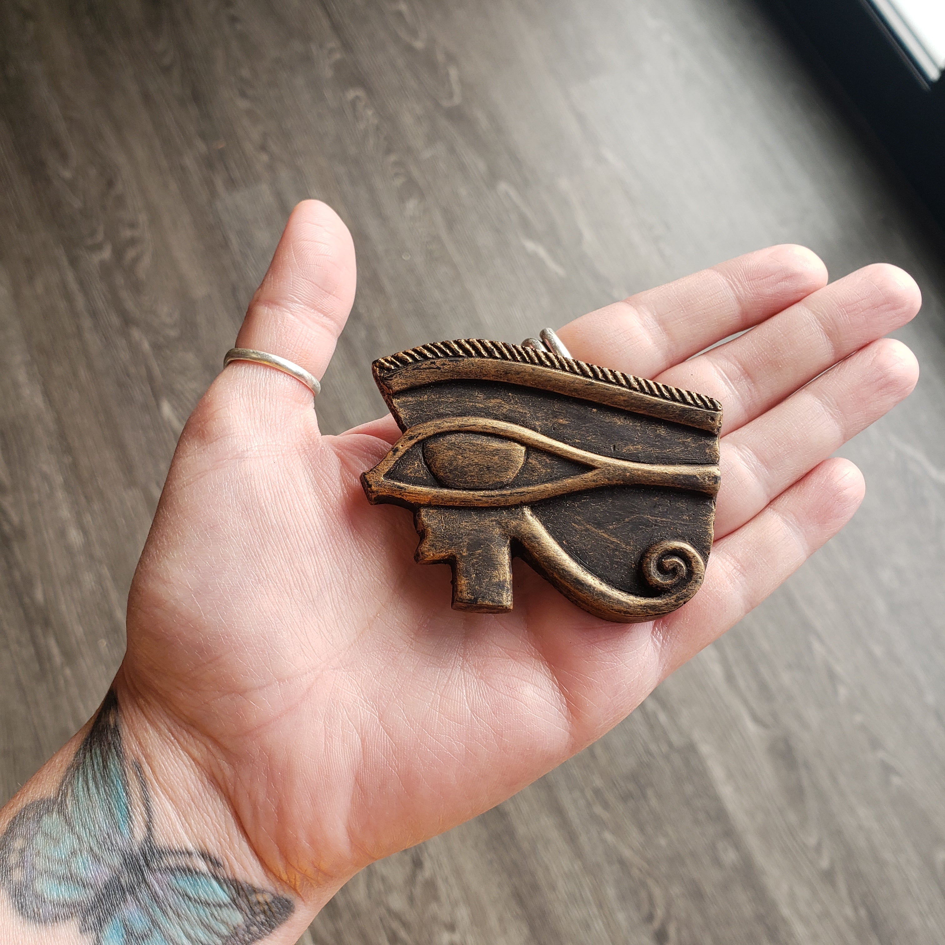 Hand holding a decorative bronze eye pendant on a wooden surface