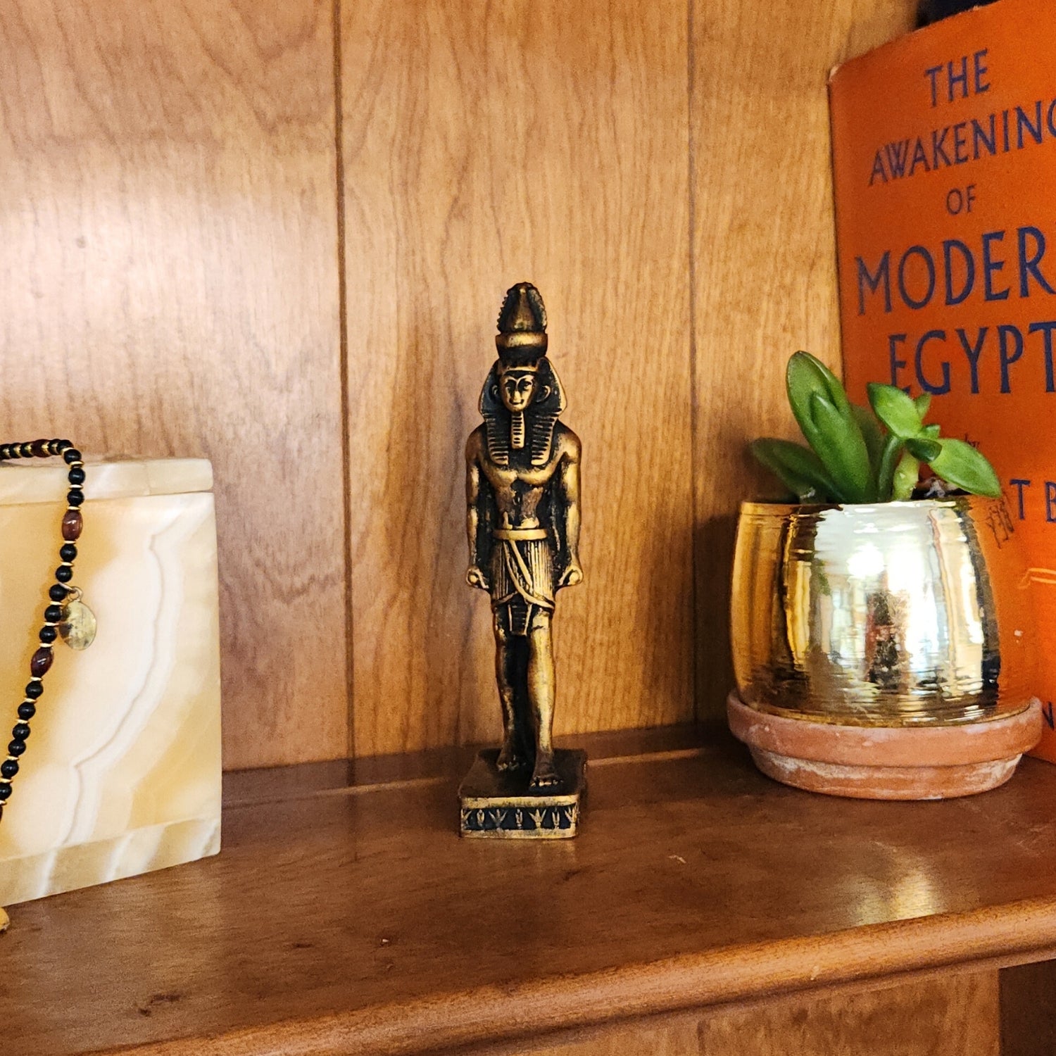 Small Egyptian-style statue on a wooden surface with a plant and decorative box.