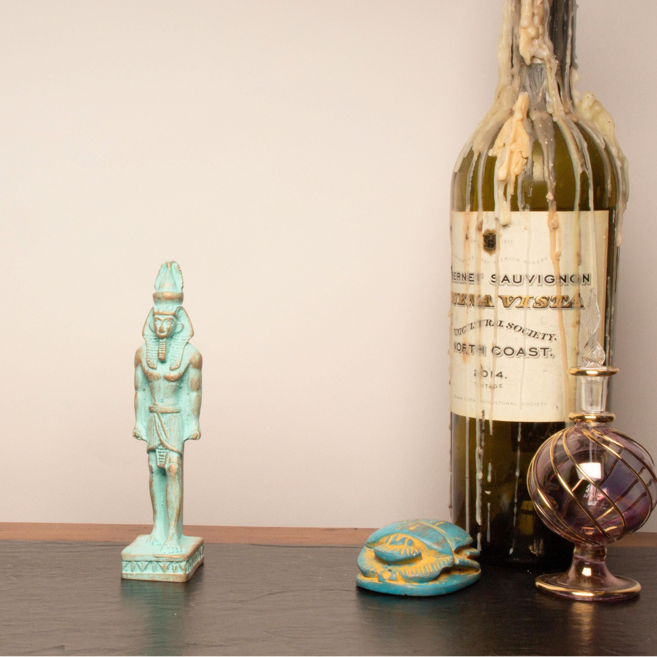 A vintage green wine bottle with melted wax dripping from the neck, styled on a dark wooden surface alongside a turquoise Egyptian pharaoh figurine, a small turquoise and gold turtle figurine, and a purple glass perfume bottle against a plain beige wall.