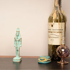 A vintage green wine bottle with melted wax dripping from the neck, styled on a dark wooden surface alongside a turquoise Egyptian pharaoh figurine, a small turquoise and gold turtle figurine, and a purple glass perfume bottle against a plain beige wall.