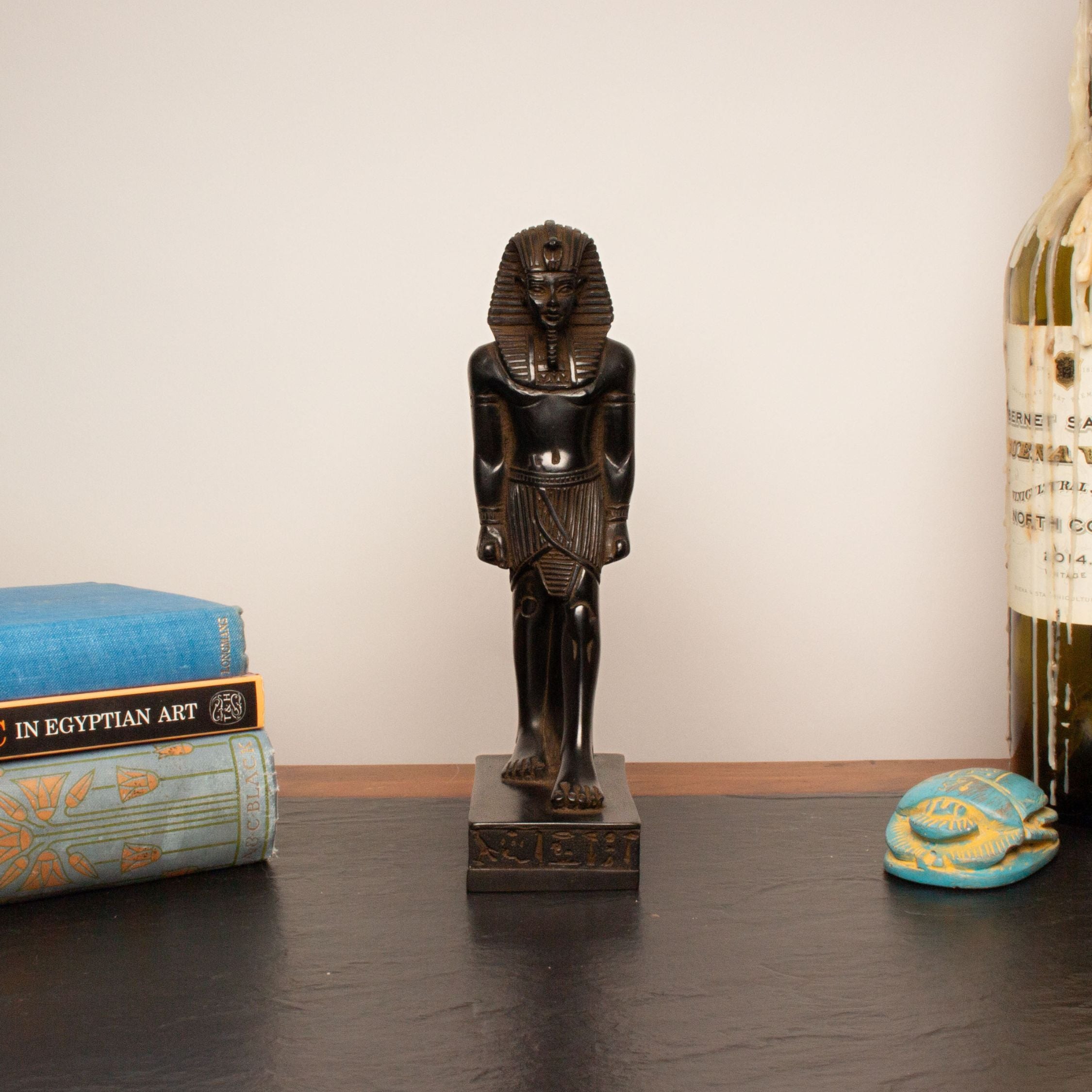 A small dark brown carved Egyptian pharaoh statue standing on a rectangular base, displayed on a dark tabletop with books and a bottle in the background.
