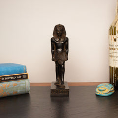A small dark brown carved Egyptian pharaoh statue standing on a rectangular base, displayed on a dark tabletop with books and a bottle in the background.