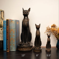 Three cat statues of different sizes on a wooden surface with books and decorative items.