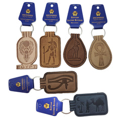 Four Egyptian leather keychains and two faux leather keychains with blue 'Discoveries Egyptian Leather Keychains' tags, featuring embossed designs of a scarab beetle, Anubis, a cat, an Ankh, an Eye of Horus, and camels on a white background.