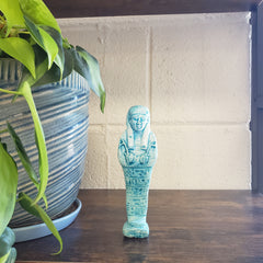 Blue Egyptian ushabti statue on a shelf with plants in the foreground