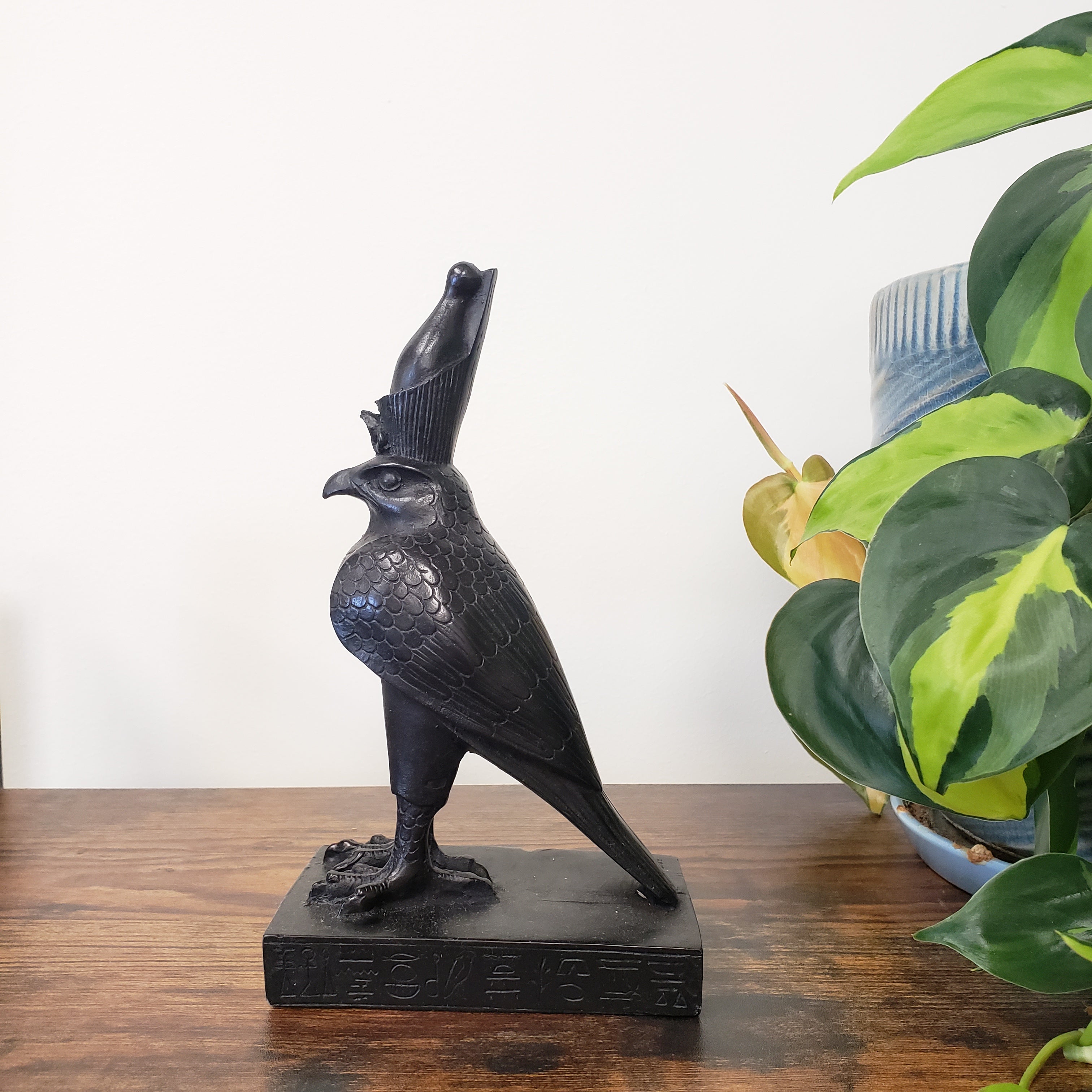 Black bird-shaped Egyptian falcon-god Horus statue on a wooden surface with green leaves in the background