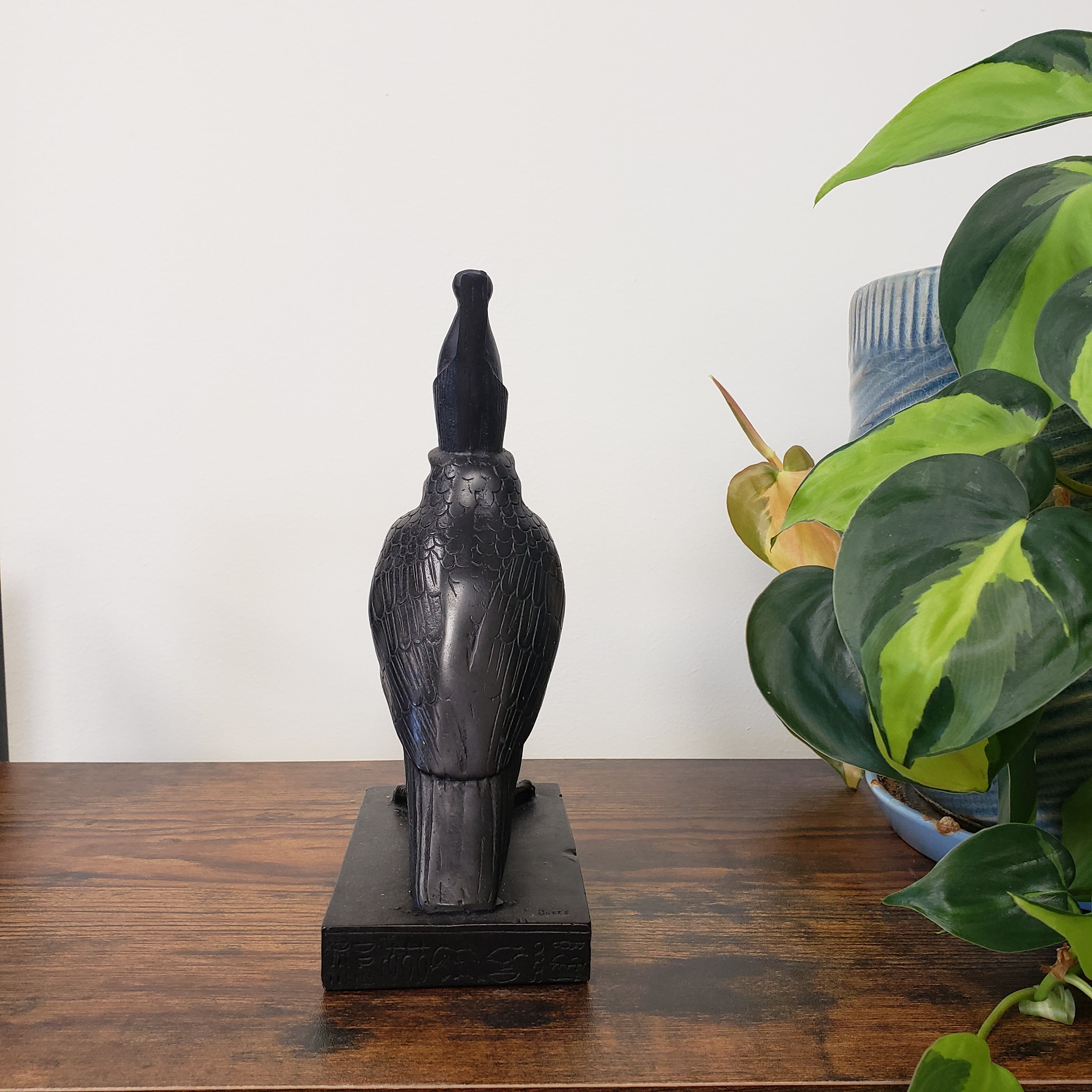 Black Egyptian falcon-god Horus sculpture on a wooden surface with green plants in the background