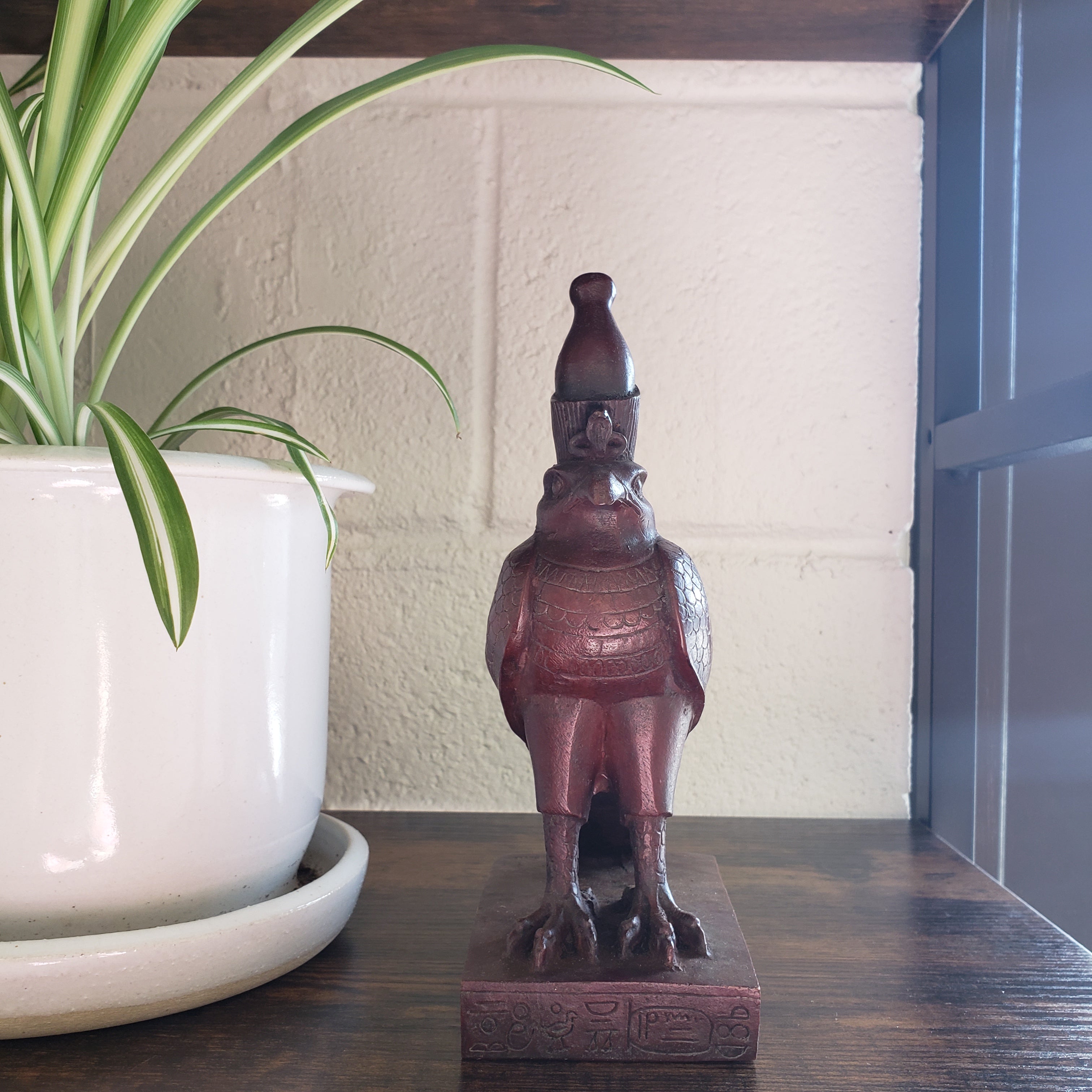 Decorative Egyptian falcon-god Horus figurine on a shelf with a plant and white container in the foreground.
