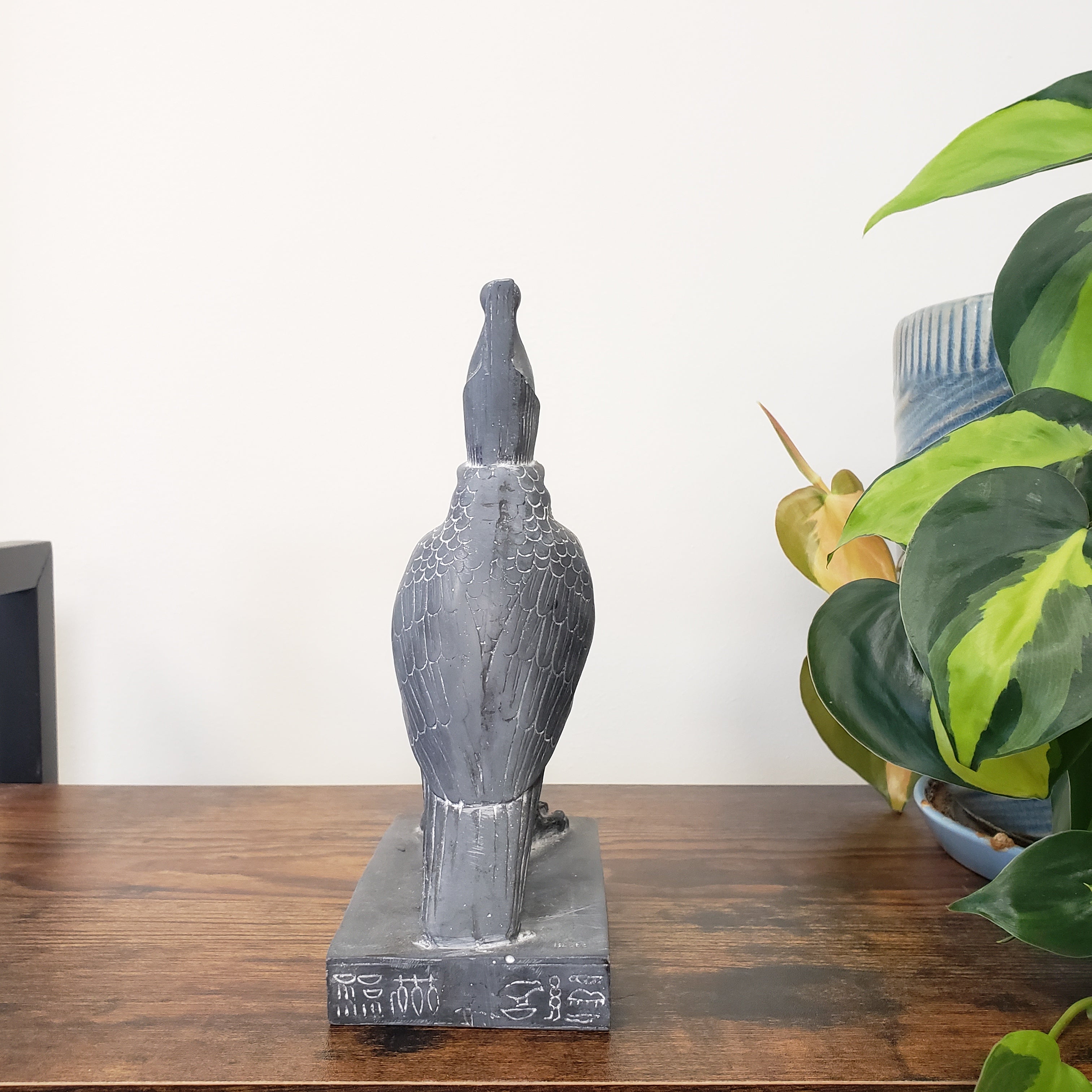 Decorative statue of a winged Egyptian falcon-god Horus on a wooden surface with a plant in the background